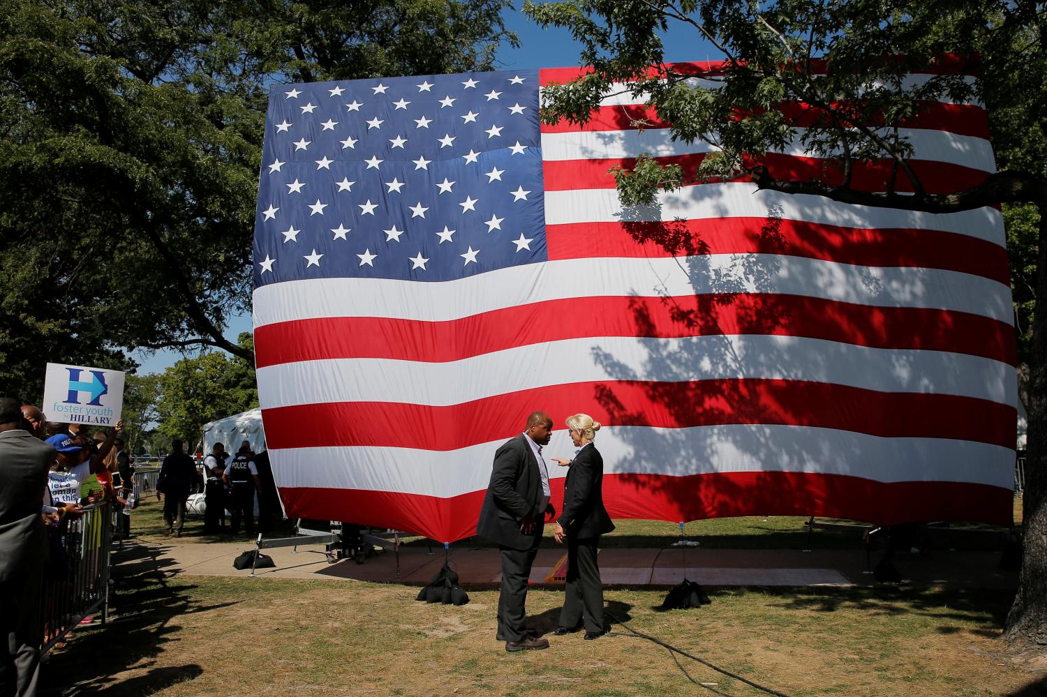 Two secret service agents, a man and a woman, talk in front of a large American flag.