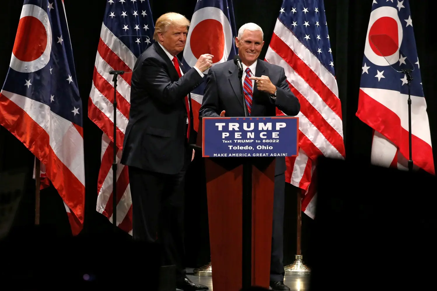 At an event in Toledo, Ohio, Mike Pence stands at the podium next to Donald Trump, and both point at the other while smiling. In the background are alternating U.S. and Ohio flags.