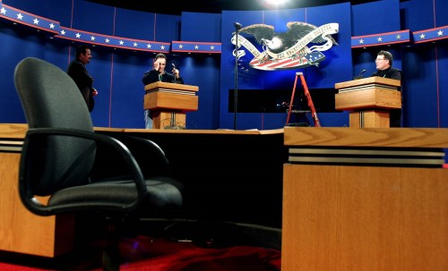 Two production staff stand at the debate podiums while the stage is being prepped. A ladder stands in the background.