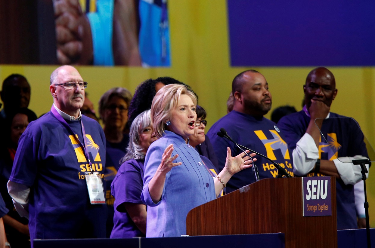 Hillary Clinton speaks at the podium of an SEIU event, joined by SEIU members in matching t-shirts.