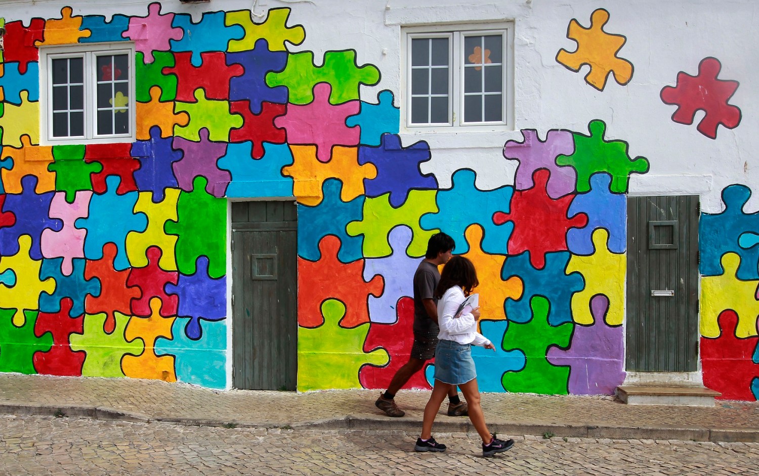 People walk past a house painted in a puzzle pattern in Eiriceira village North of Lisbon August 13, 2011. Portugal still faces some tough challenges but is on track to meet this year's budget deficit goal despite a shortfall in performance so far, officials from the European Union and IMF said on Friday. REUTERS/Jose Manuel Ribeiro (PORTUGAL - Tags: POLITICS BUSINESS)