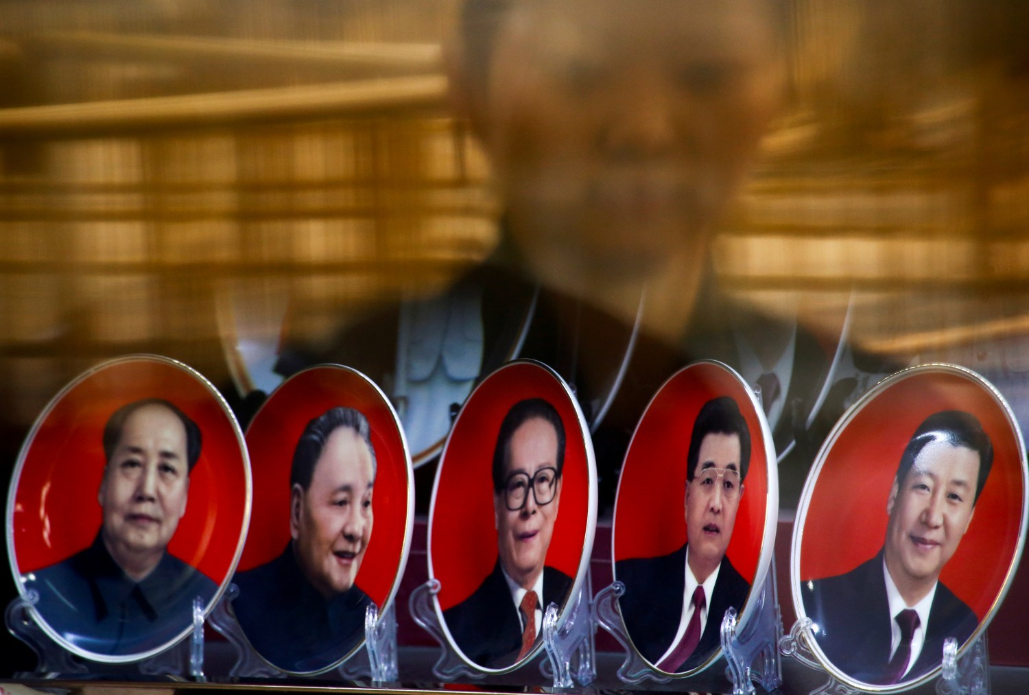 A woman is reflected in a shop window as she looks at souvenir plates with portraits of former Chinese leaders Mao Zedong, Deng Xiaoping, Jiang Zemin, Hu Jintao and current President Xi Jinping