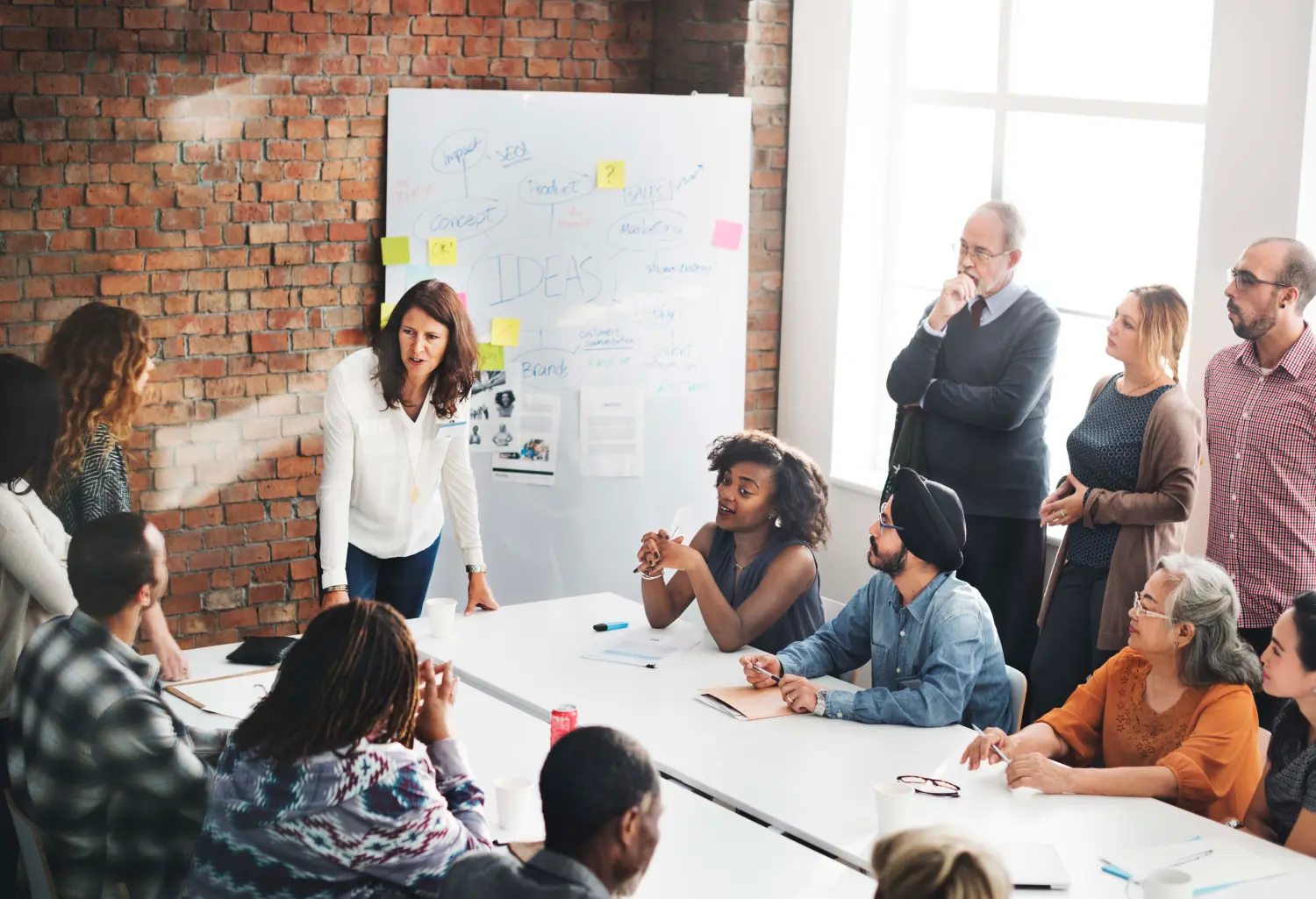 An older woman stands at the head of a table of mixed-age workers in a modern office space [photo courtesy of iStock]