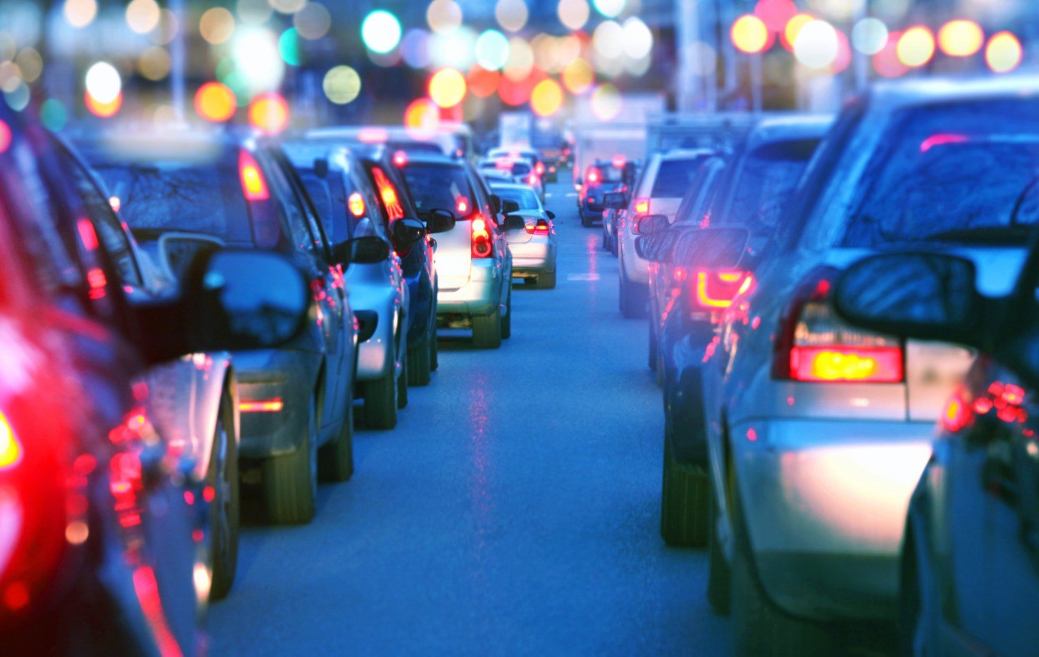 iStock Images. Driving a car in a a city with a traffic jam at night, proceeding slow in a line of cars with red tail lights during the rush hour. view from inside the car. Blurred streets lights on the background. Dusk, blue skylight.