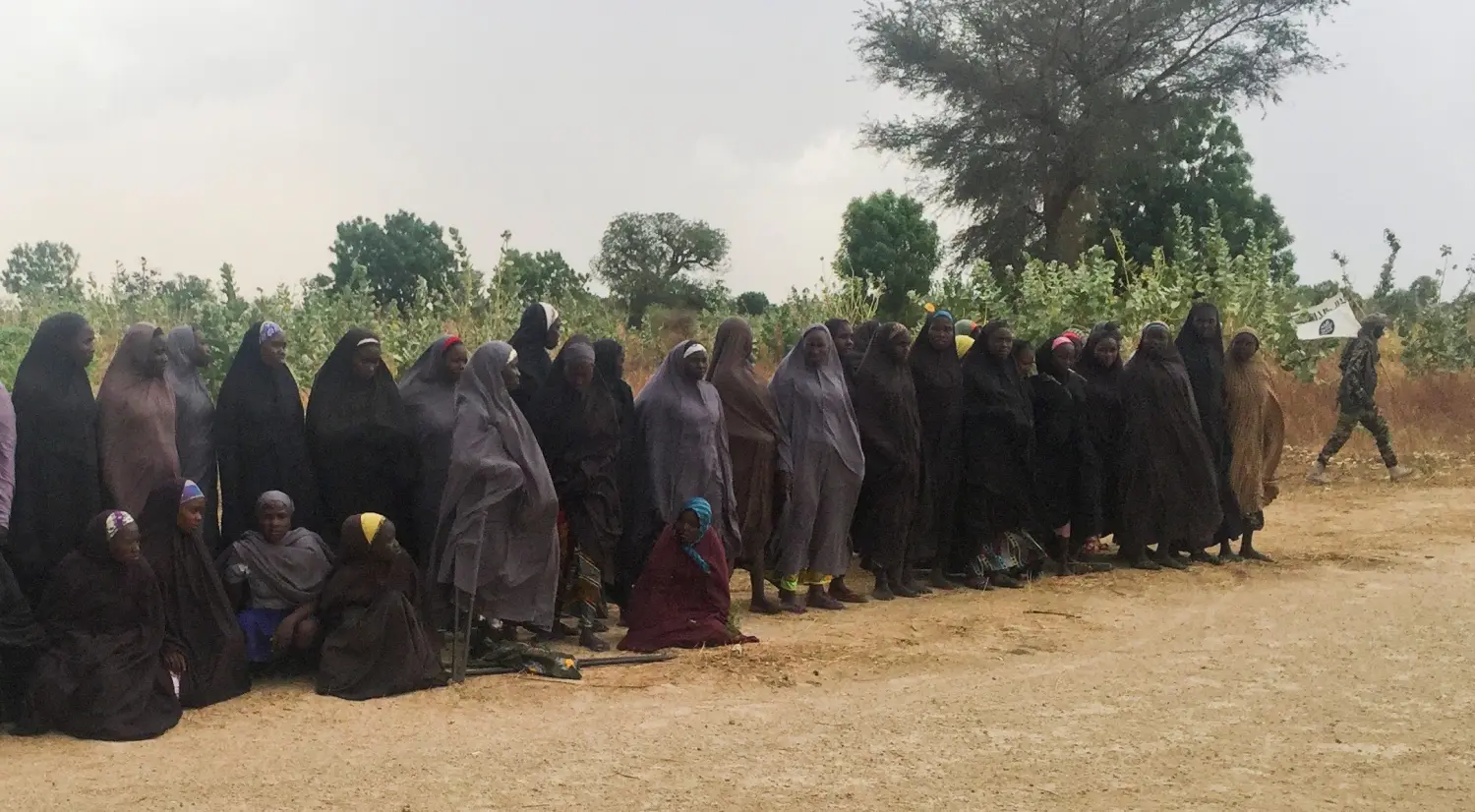 A man carrying a Boko Haram flag walks past a group of 82 Chibok girls, who were held captive for three years by Islamist militants, as the girls wait to be released in exchange for several militant commanders, near Kumshe, Nigeria May 6, 2017. Picture taken May 6, 2017. REUTERS/Zanah Mustapha - RTS15P0S