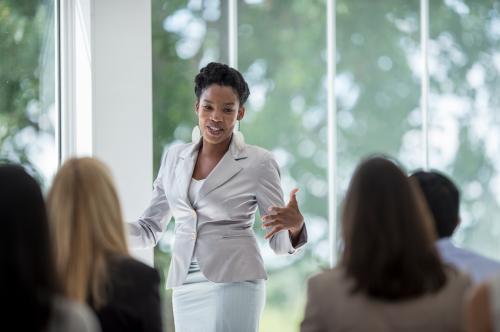 A multi-ethnic group of business professionals are getting training at the corporate office. A woman is presenting on their business concepts.