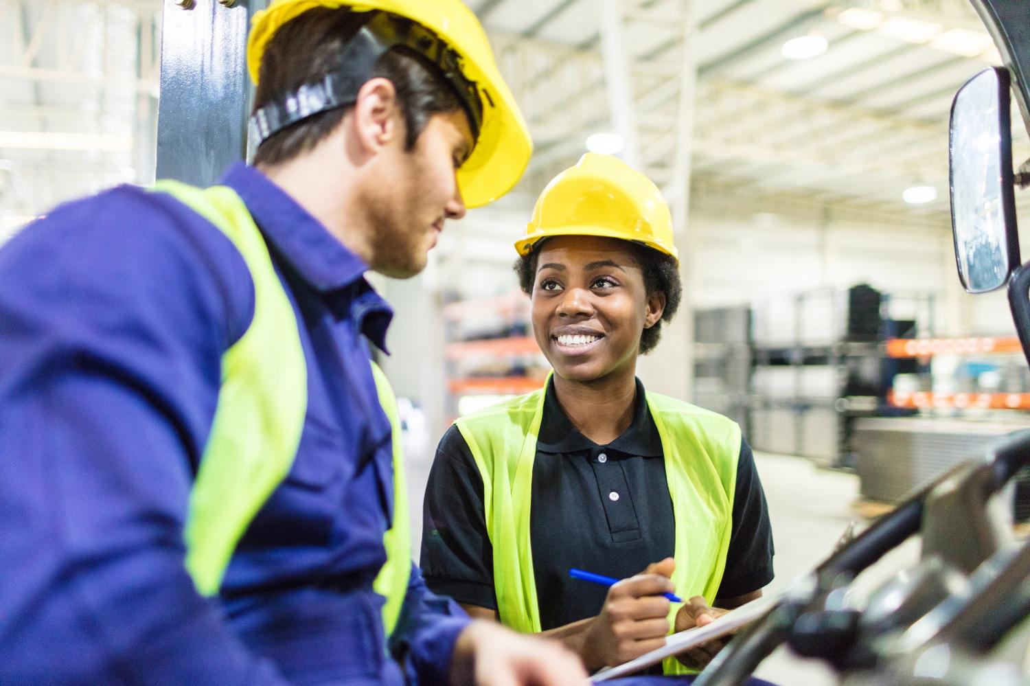 Woman talking to forklift truck operator in warehouse. Warehouse workers discussing shipping schedule.