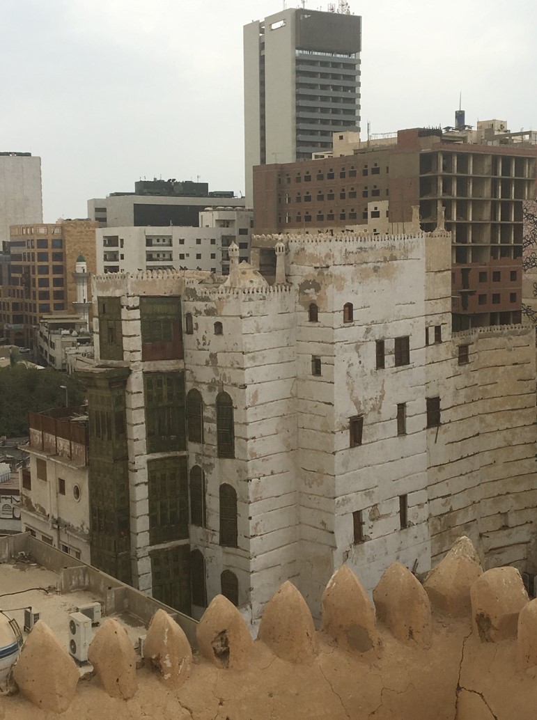 Looking from the roof of Al Nassif House across to another historical multistory building, with modern Jeddah crowding in behind. Kitchens were built on the top floor—thus you can see the dome and chimneys of the white home’s kitchen directly in front of you. The horizontal lines on the house are dark wood planks, placed between layers of mud brick to support the multiple stories.