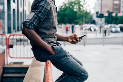 From the neck down view of young afro black man holding a smart phone, tapping the scree outdoor in the city - technology, social network, communication concept