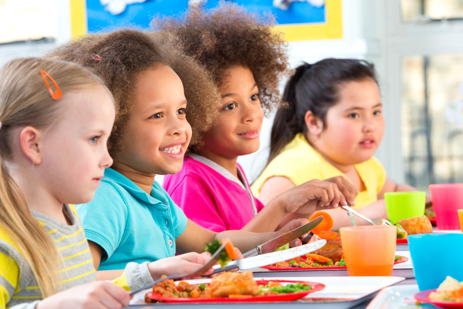 Four children can be seen at a school dinner table with meals infront of them. They are looking at something and smiling, with their cutlery in their hands.