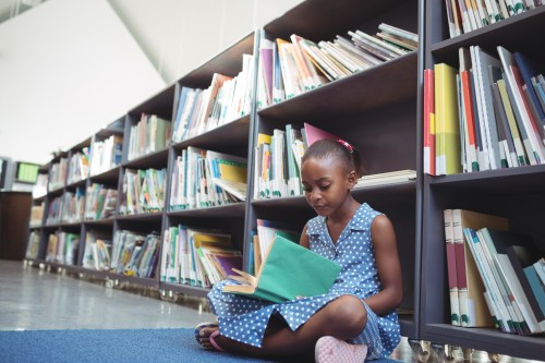 Girl reading book while sitting by shelf in library