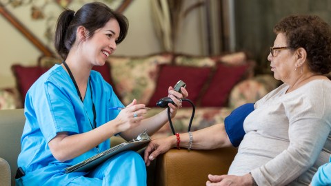 Cheerful Asian nurse checks Hispanic senior woman's blood pressure in nursing home. The nurse is holding the blood pressure gauge. She is wearing blue scrubs.