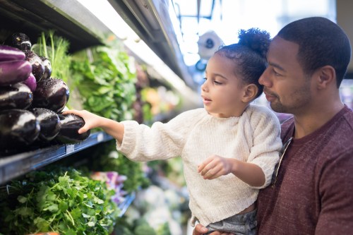 A pre-school age girl helps her dad pick out veggies in the produce section at the grocery store. He is holding her next to the produce and she is picking out eggplant.