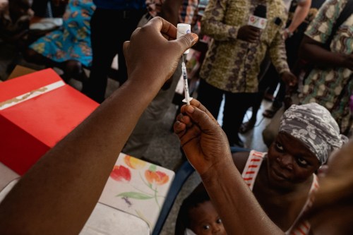 A nurse holds a syringe as patients wait in Ghana April 23, 2019. Gavi/2019/Tony Noel. Picture taken April 23, 2019. Zipline drones, supported by Gavi and the UPS Foundation, cut the time taken to deliver lifesaving medical supplies from hours to minutes. Gavi/2019/Tony Noel via REUTERS   ATTENTION EDITORS - THIS IMAGE HAS BEEN SUPPLIED BY A THIRD PARTY. MANDATORY CREDIT - RC187D079370