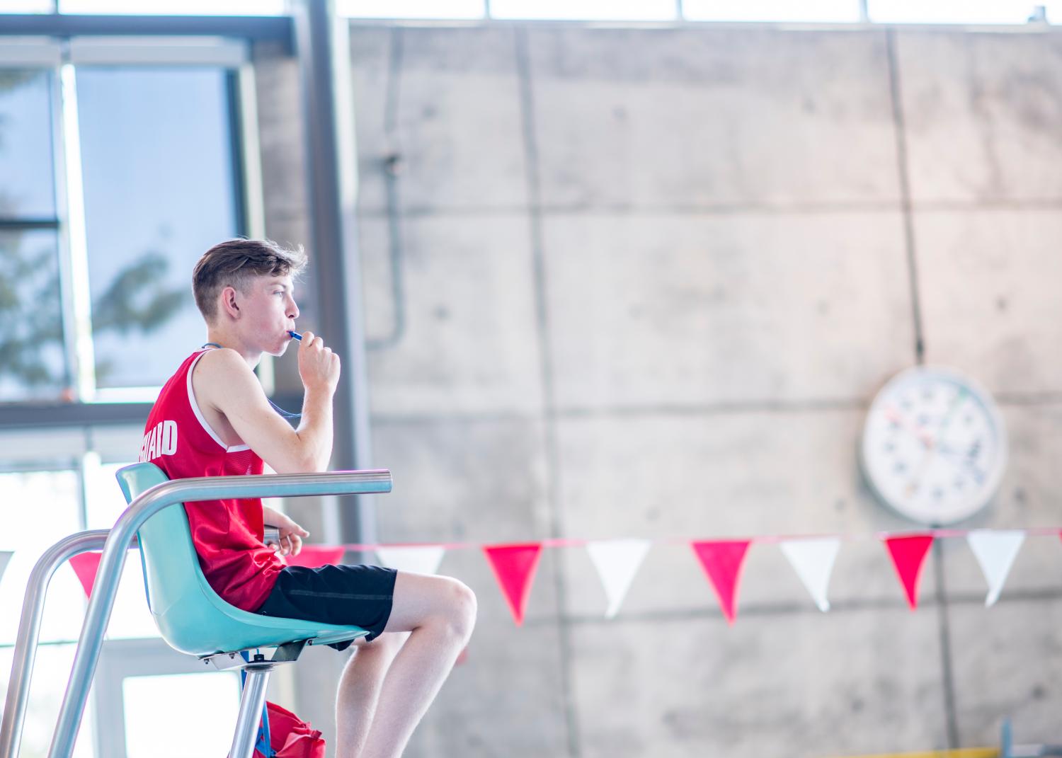 A teen lifeguard is sitting and watching over the pool. He is about to blow his whistle.