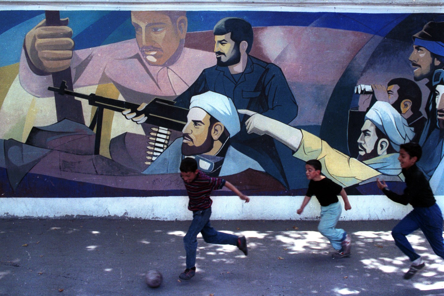 IRANIAN CHILDREN PLAY SOCCER BY HUGE GRAFFITI SHOWING MULLAHS LEADING REVOLUTIONARY GUARD TO WAR AGAINST IRAQ.  Iranian children play soccer by a huge grafitti showing mullahs leading the revolutionary guard to the war against Iraq in Tehran 6 June, 1990. SCANNED FROM NEGATIVE REUTERS/Yannis Behrakis - RP1DRICQVKAD