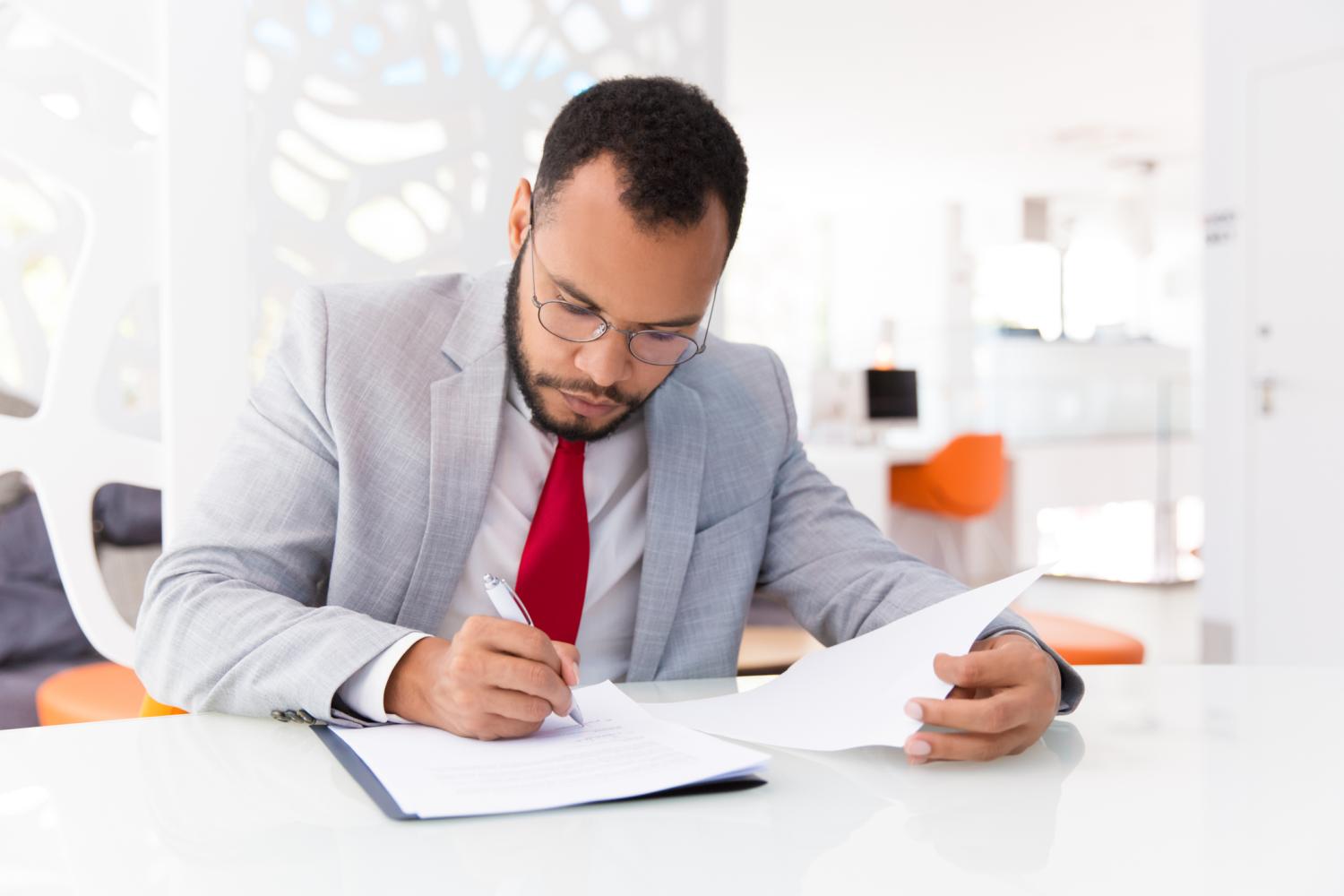 Focused auditor checking document. Business man wearing suit and eyeglasses, signing contract. Document expertise concept