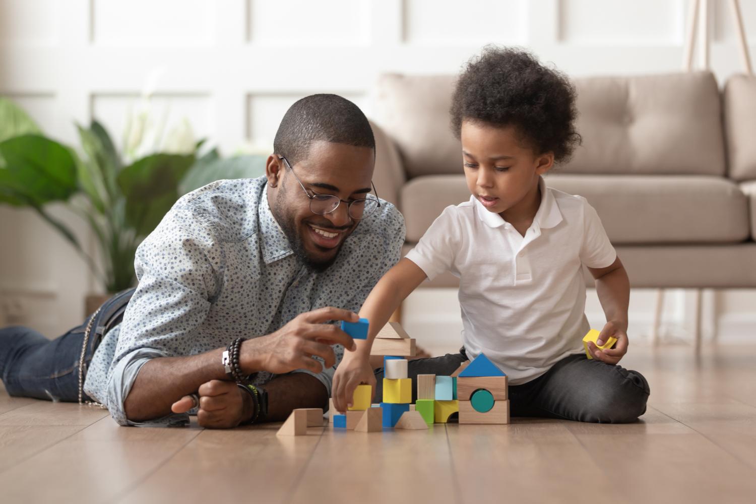 Caring young single black father help cute kid son play on warm floor together, happy african family dad and little child boy having fun building constructor tower from colorful wooden blocks