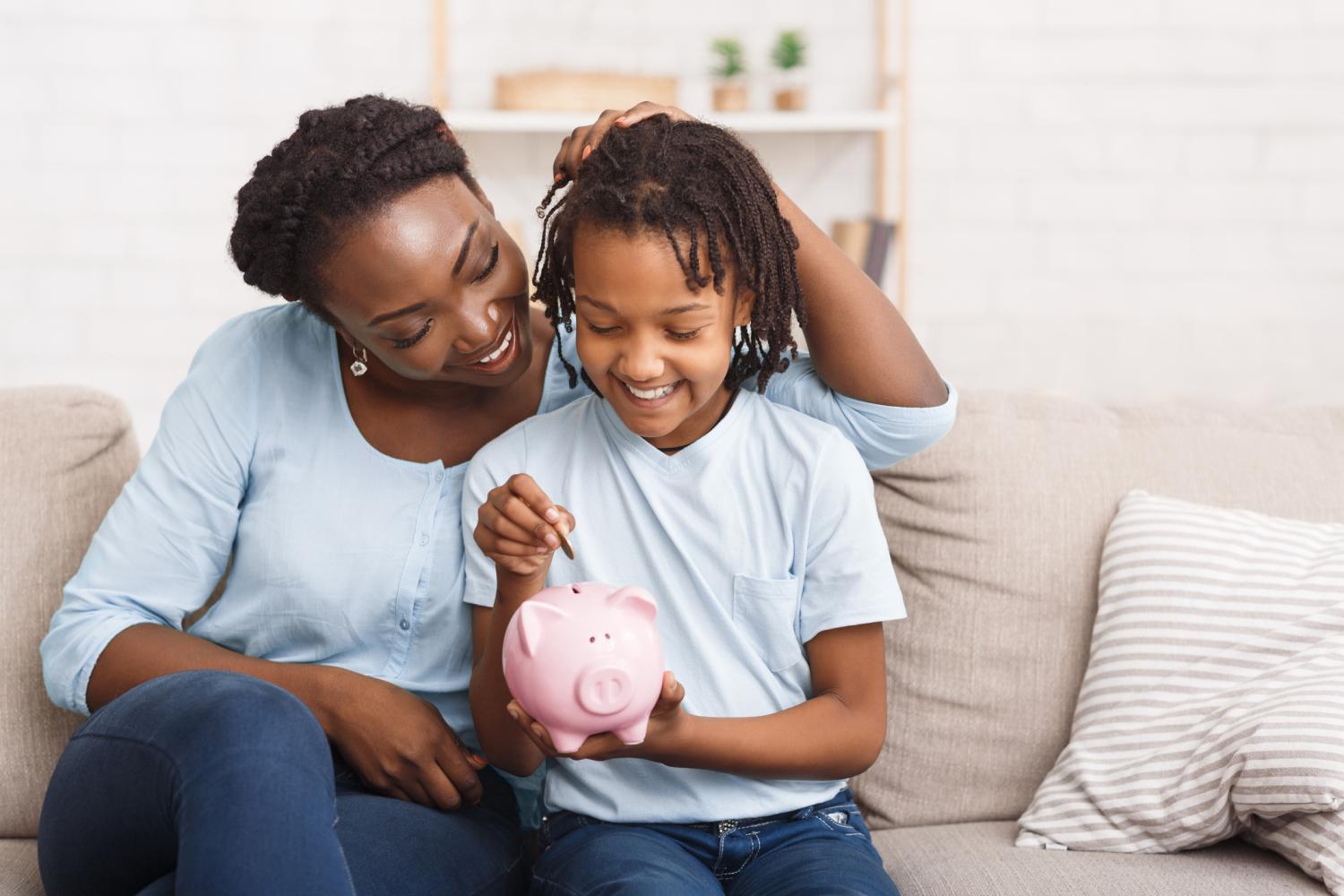 Black Mother And Daughter Putting Coins Into Piggy Bank At Home.
