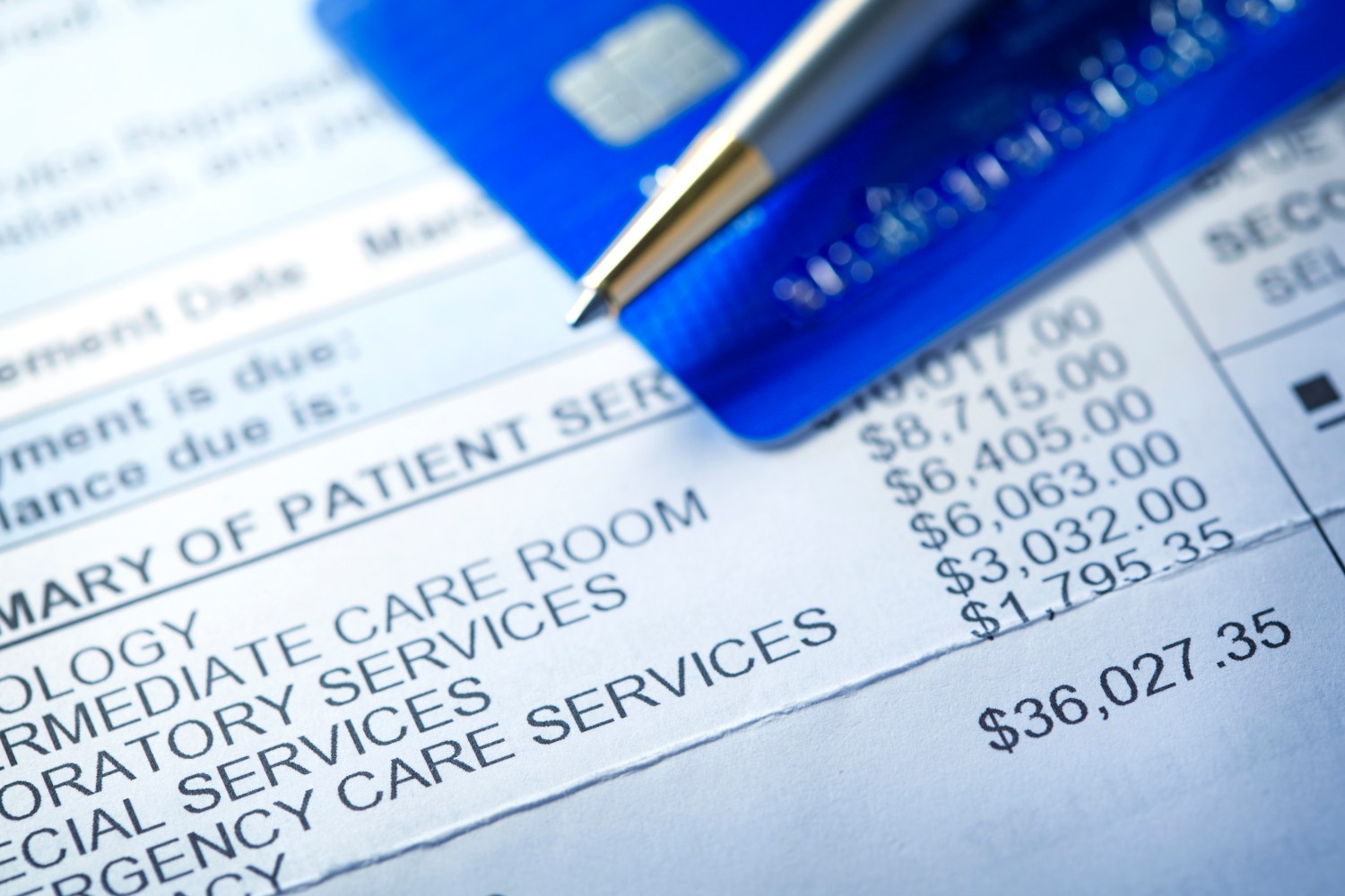A hospital bill for $36,000 with a line item of various charges is photographed with a very shallow depth of field. A credit card and a ballpoint pen rest out of focus in the background.