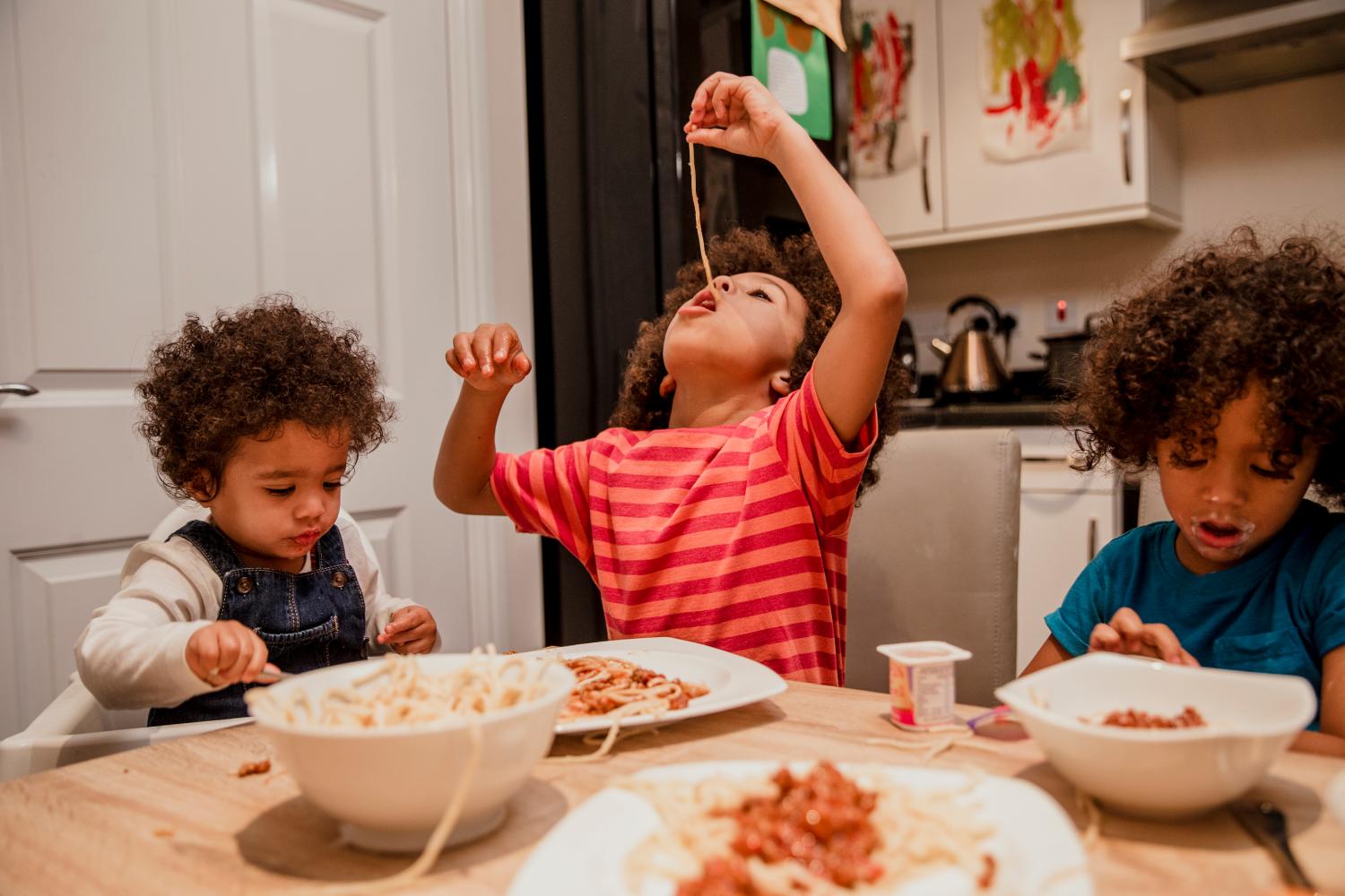 A family around a table eating spaghetti one child dangles spaghetti into his mouth