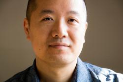 An Asian American man with closely shaved head wearing a blue, patterned collared shirt looks at the camera with a slight smile