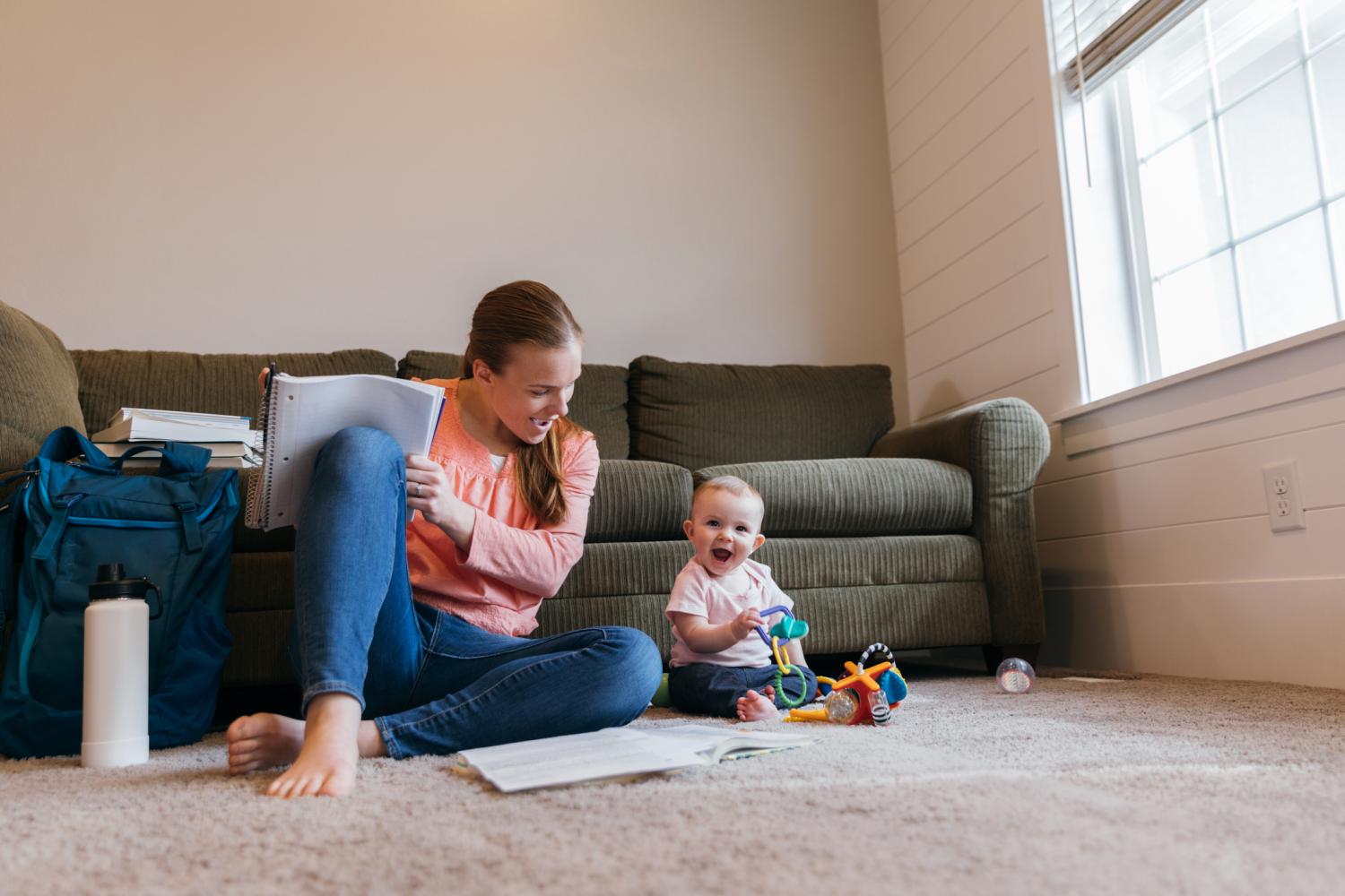 A mother and parent sits with her baby daughter on the family room floor studying for a university class. She is a hard working college student trying to earn her degree in education to improve her situation.