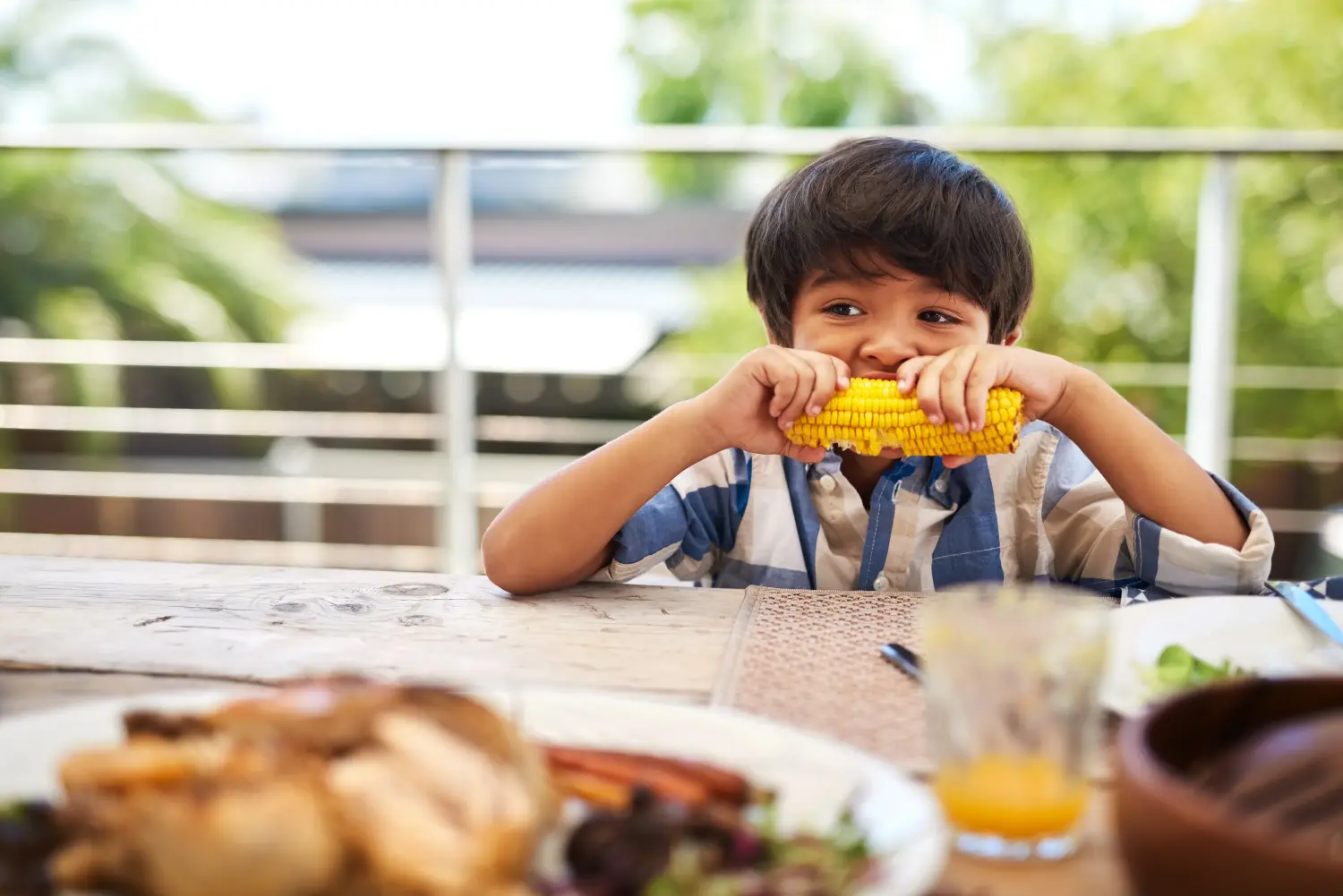 Shot of an adorable little boy eating a cob of corn around a table outdoors