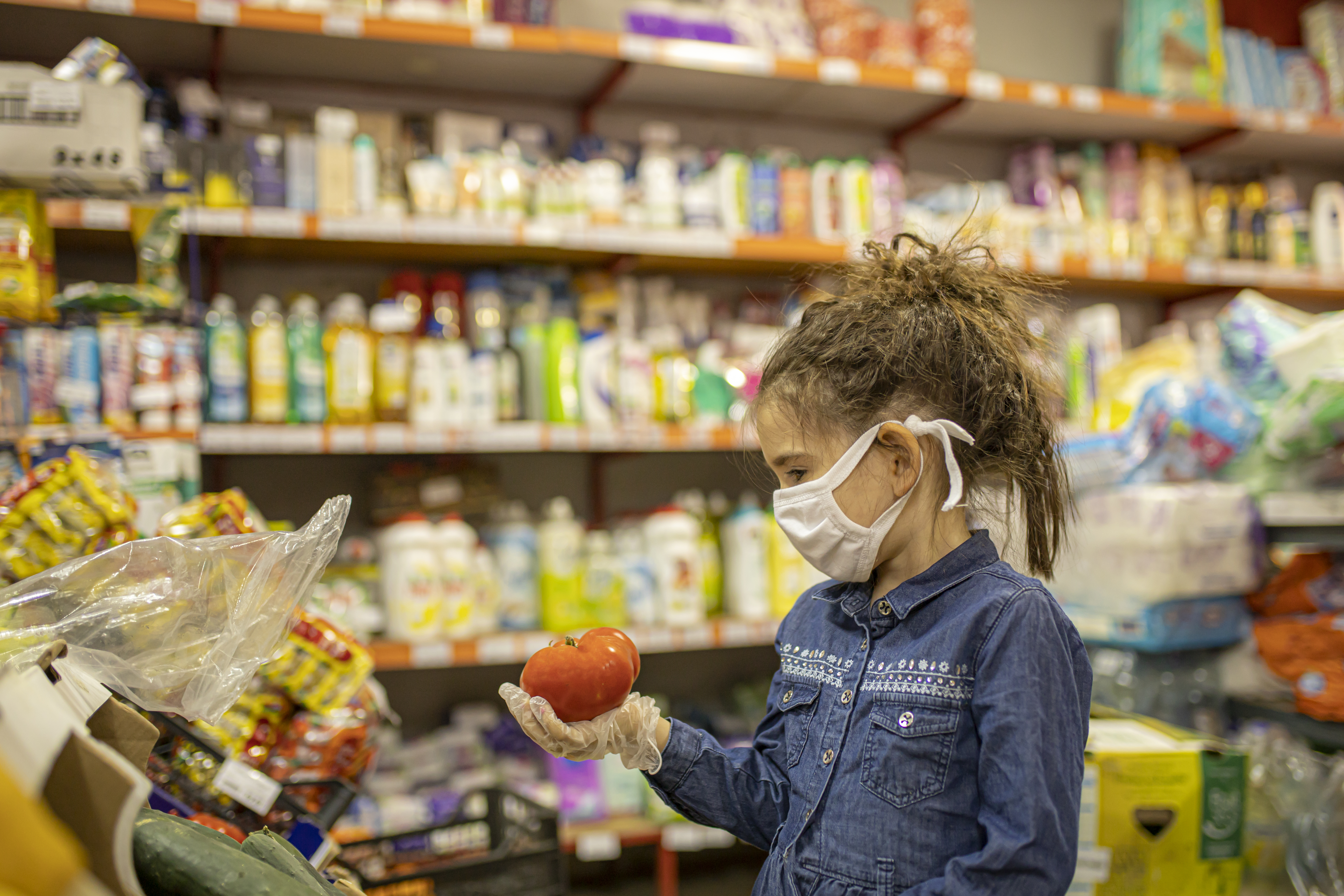 Young girl wearing face mask and gloves looking at a tomato in a grocery store.