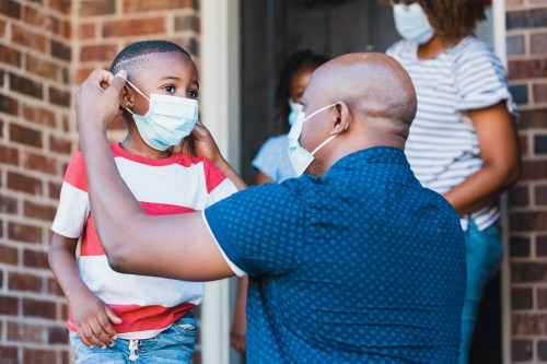 A mid adult father helps his young son put on a protective face mask before leaving their home amid the coronavirus pandemic.