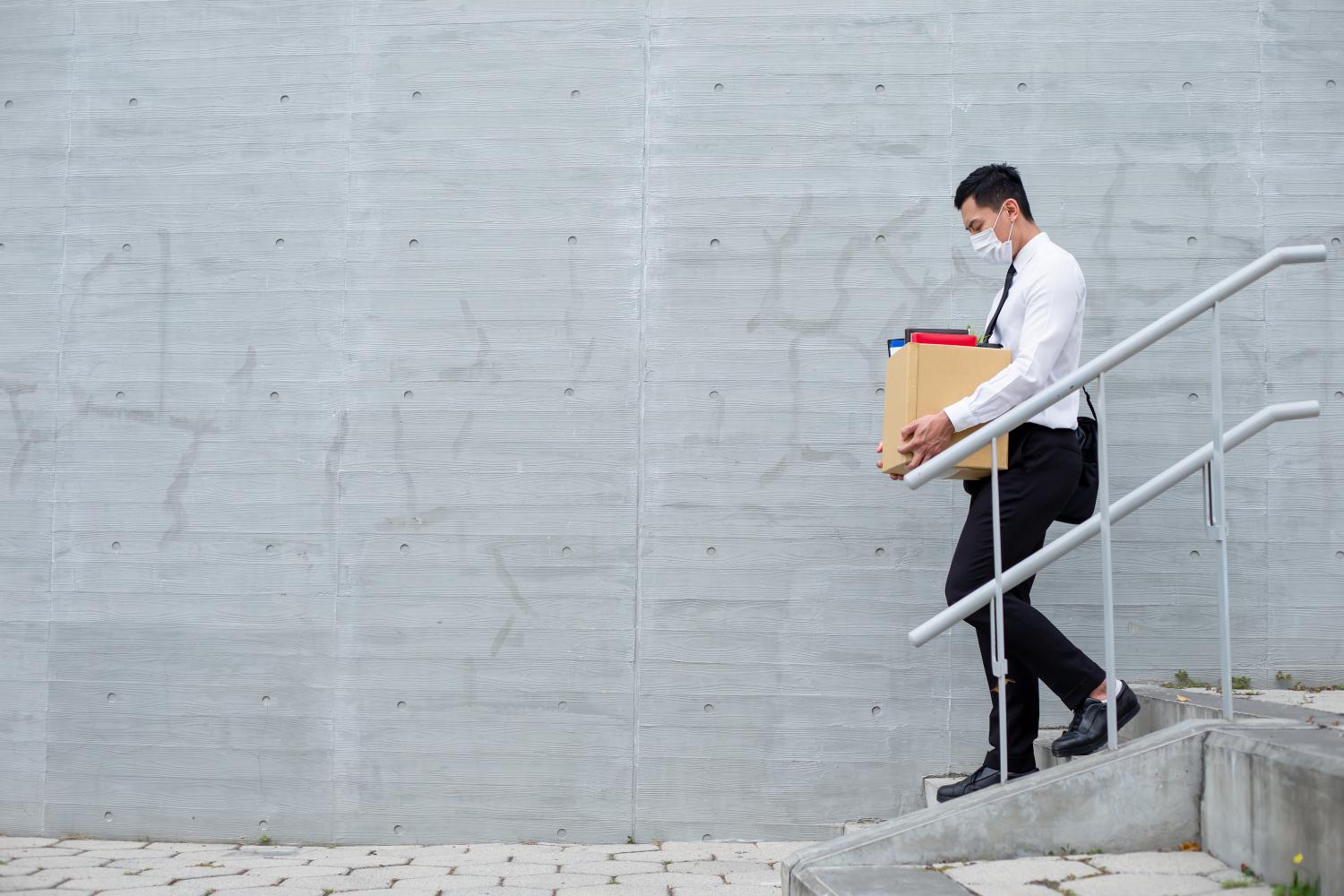 Man wearing a surgical mask carries a box of desk materials down a flight of stairs.