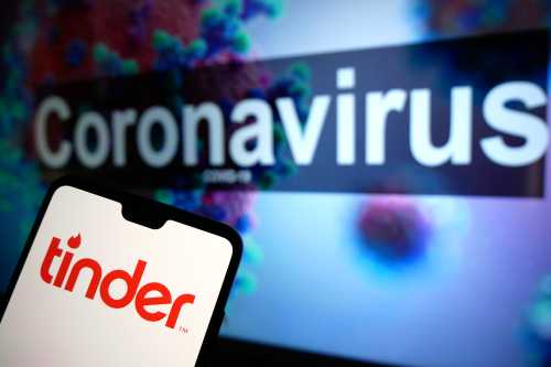 The Tinder dating app logo seen displayed on a mobile phone with an illustrative model of the Coronavirus displayed on a monitor in the background. Photo credit should read: James Warwick/EMPICS Entertainment
