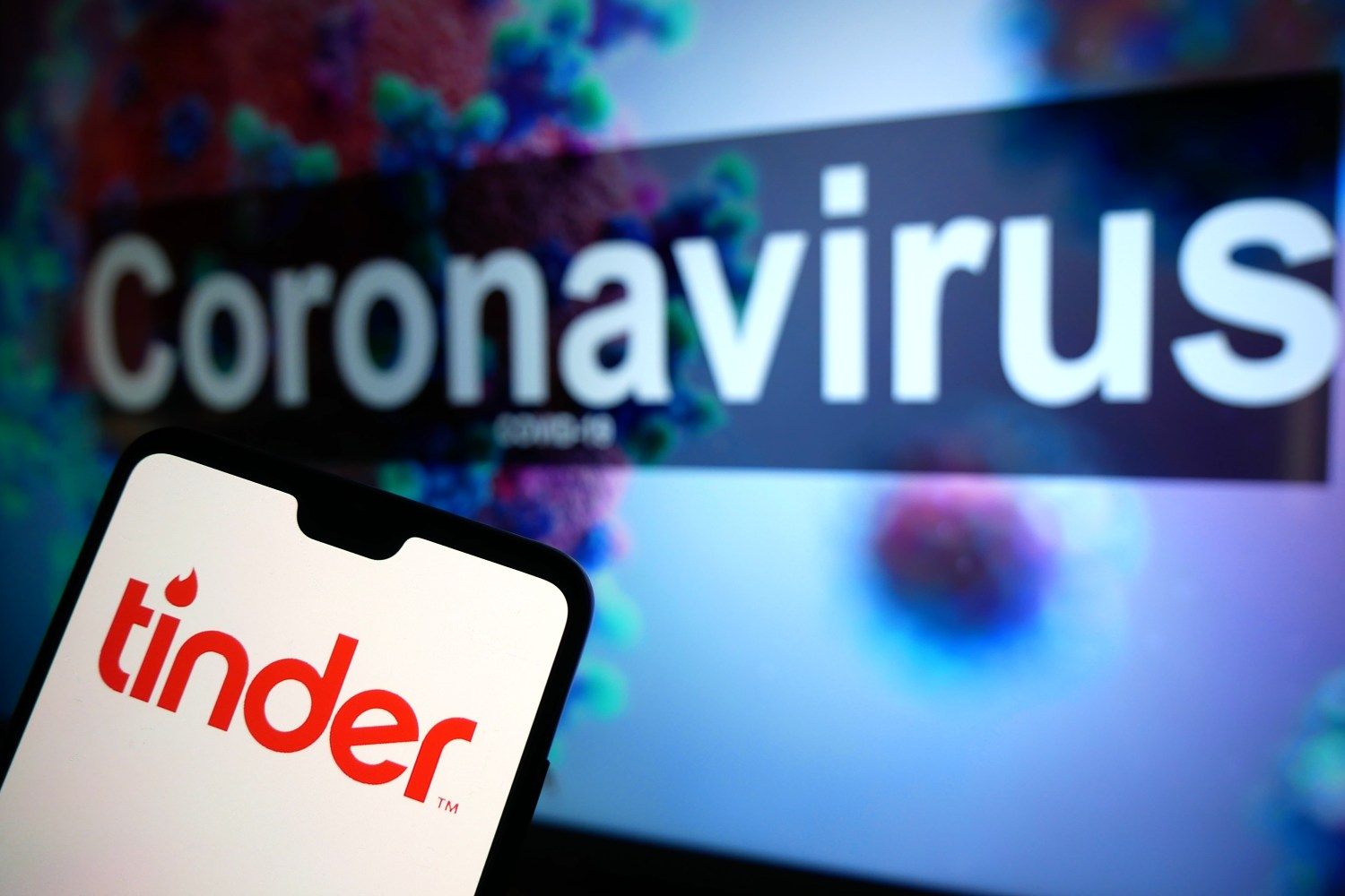 The Tinder dating app logo seen displayed on a mobile phone with an illustrative model of the Coronavirus displayed on a monitor in the background. Photo credit should read: James Warwick/EMPICS Entertainment