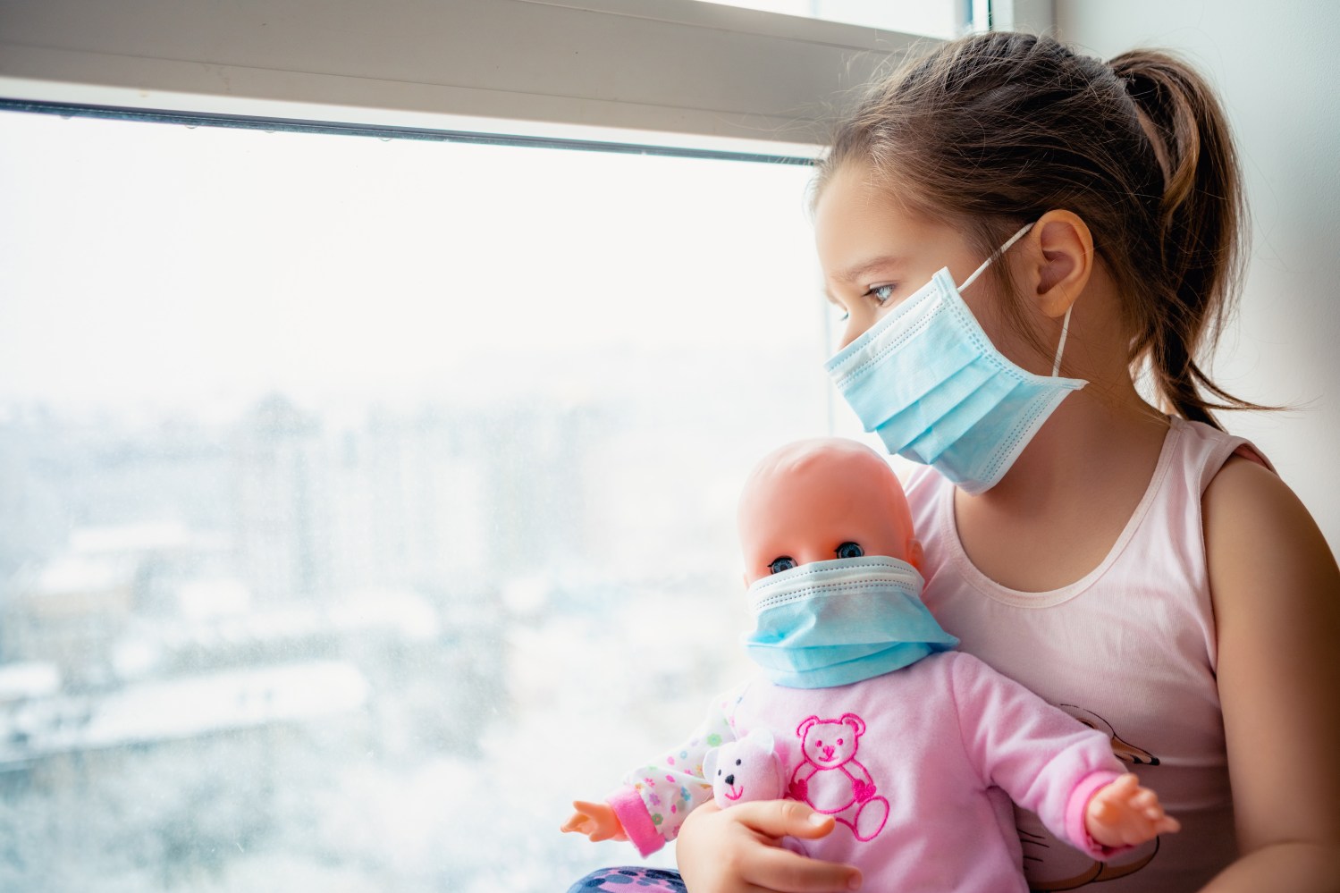 Little girl holding a doll and looking out a window wearing surgical masks.