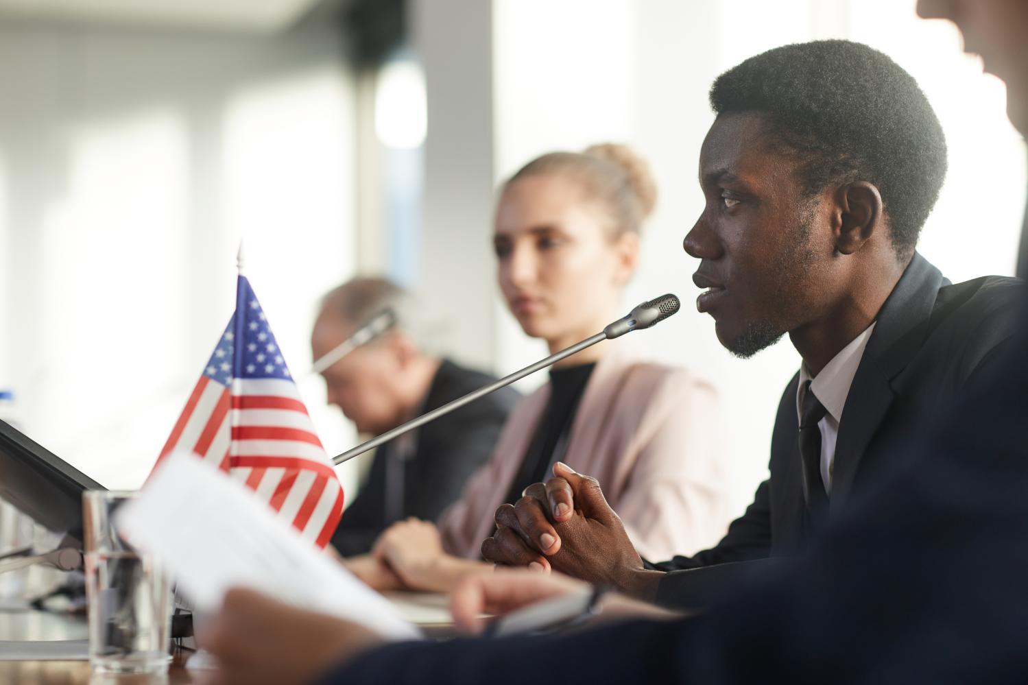 Side profile of a person speaking into a microphone with an American flag in the background.