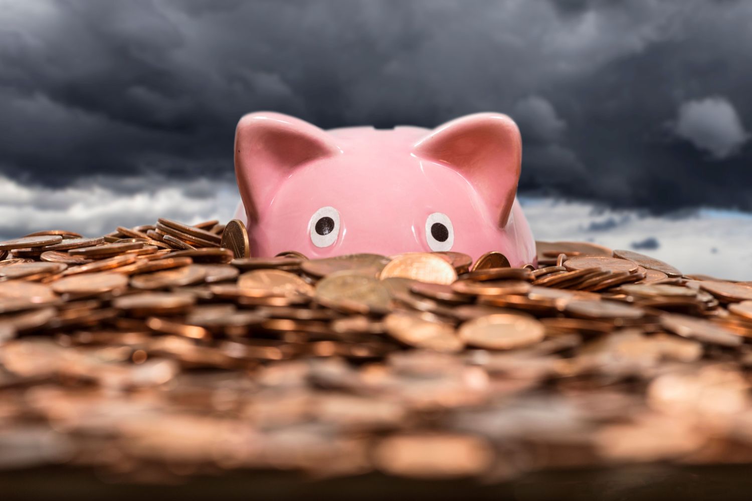 Piggy bank in an ocean of coins with gathering storm clouds in the background.