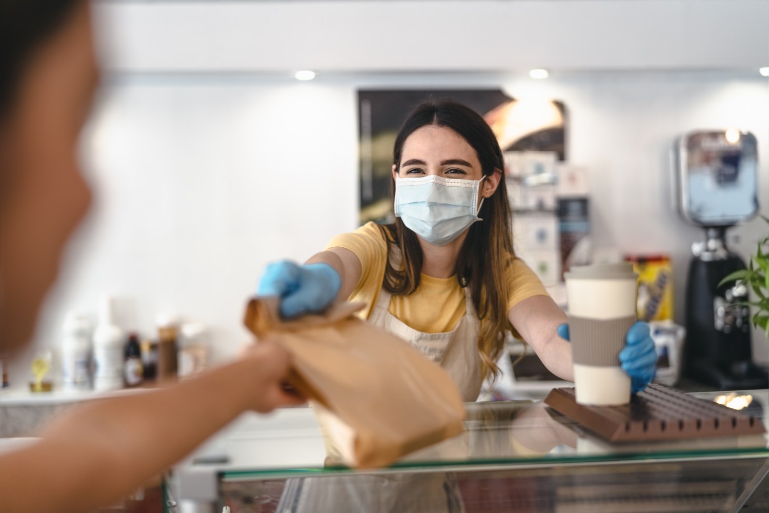 Woman wearing a surgical mask handing a customer a bag and a cup of coffee.