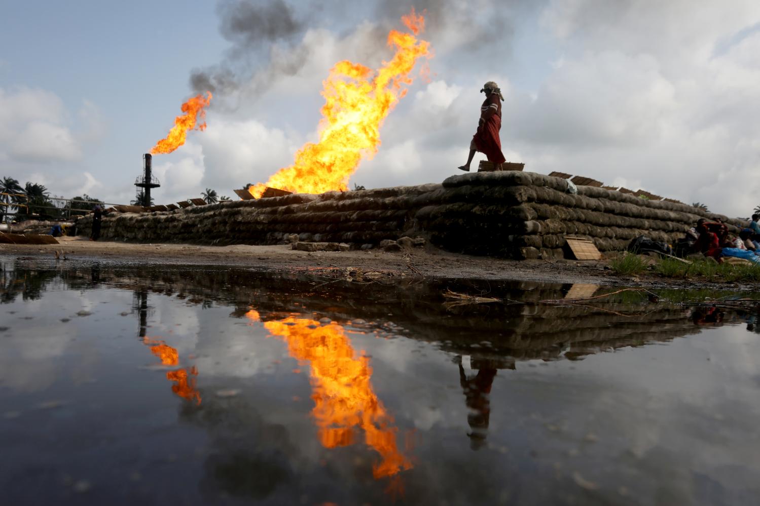 Two gas flaring furnaces and a woman walking on sand barriers are reflected in a pool of oil-smeared water at a flow station in Ughelli, Delta State, Nigeria September 17, 2020. Picture taken September 17, 2020. REUTERS/Afolabi Sotunde     TPX IMAGES OF THE DAY