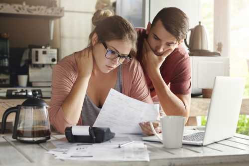 A stressed couple staring at a piece of paper by a table, which also has coffee, a laptop, and a coffee mug