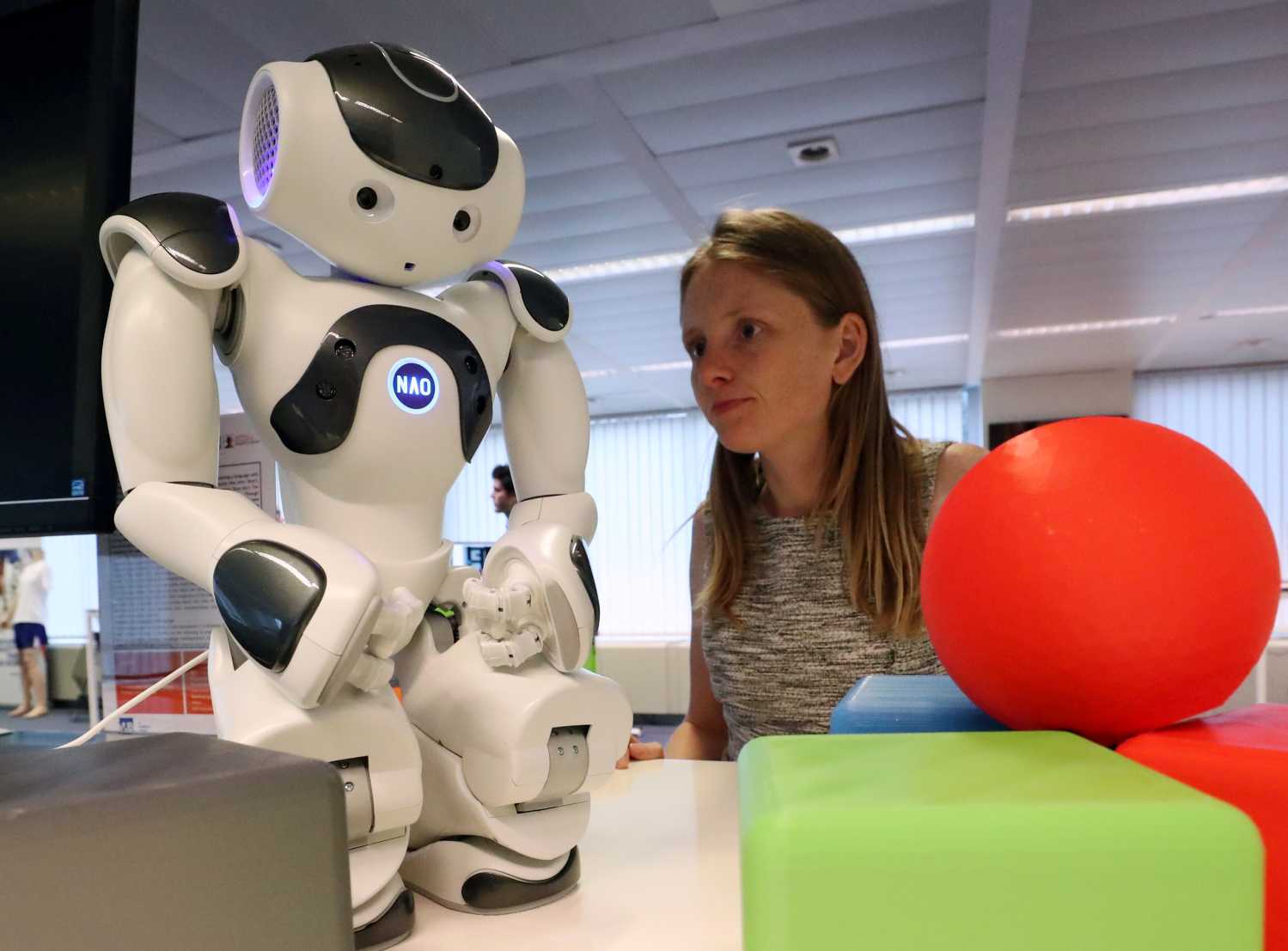 A researcher looks at a small humanoid robot standing among red and green plastic objects.