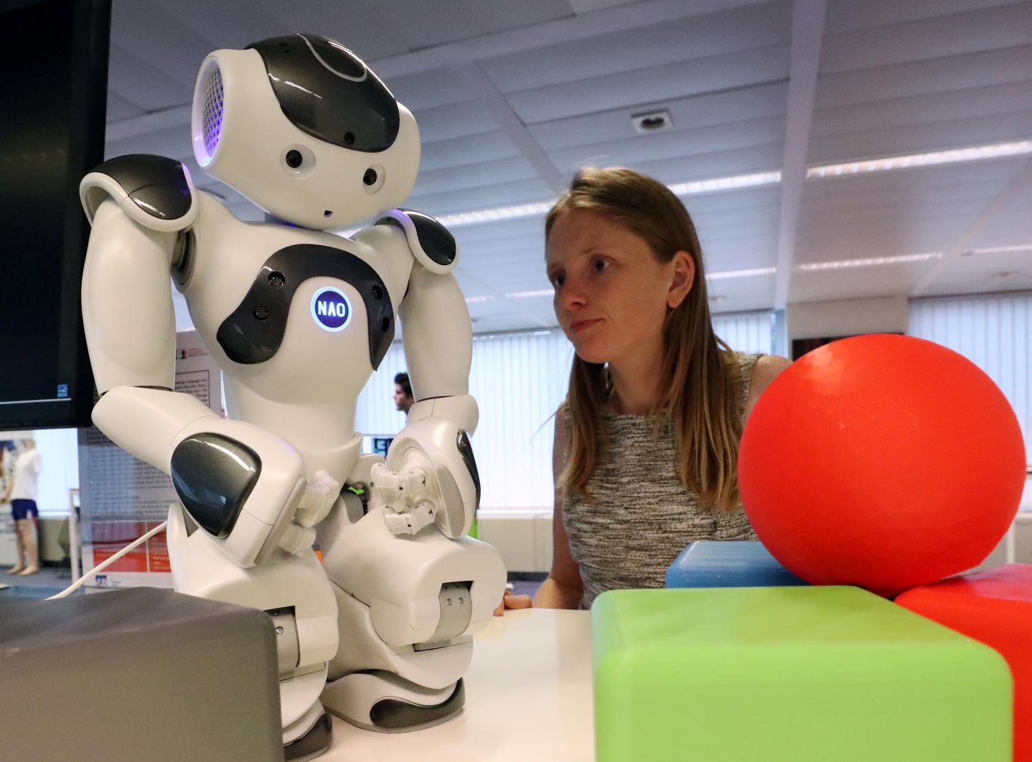 A researcher looks at a small humanoid robot standing among red and green plastic objects.
