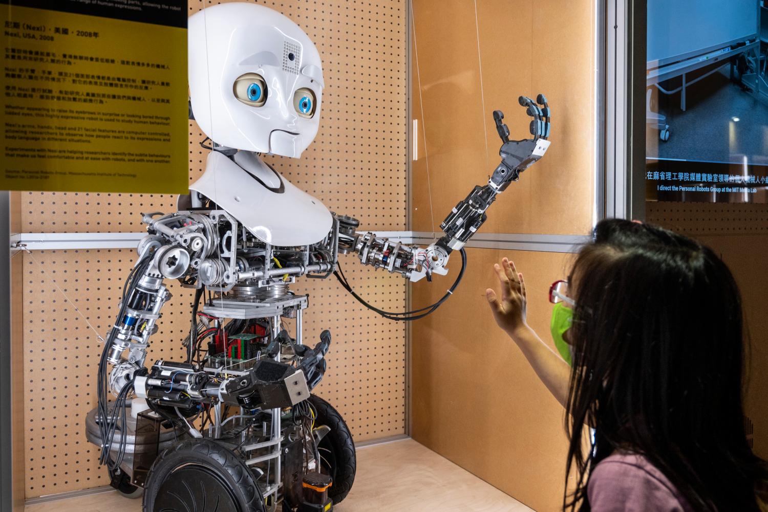 A young girl puts her hand up to a robot with a humanoid face and arm.