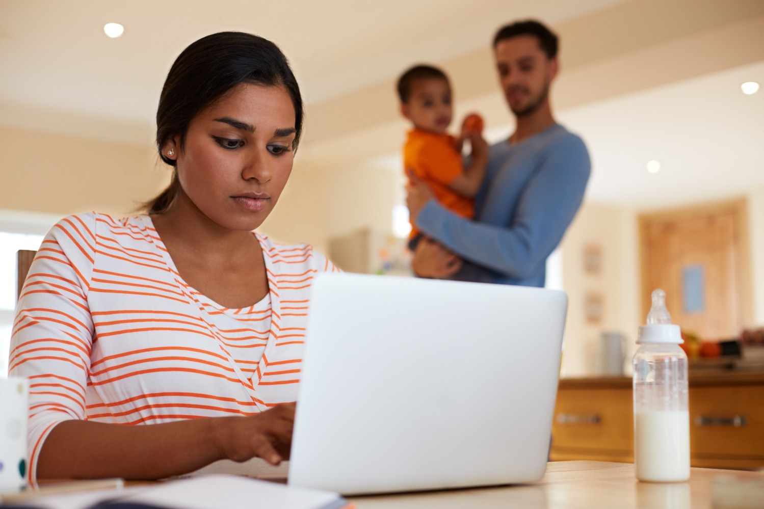 Woman working at her laptop with husband and child in the background.