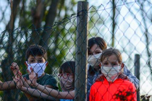 Children with cloth face masks standing behind a fence.