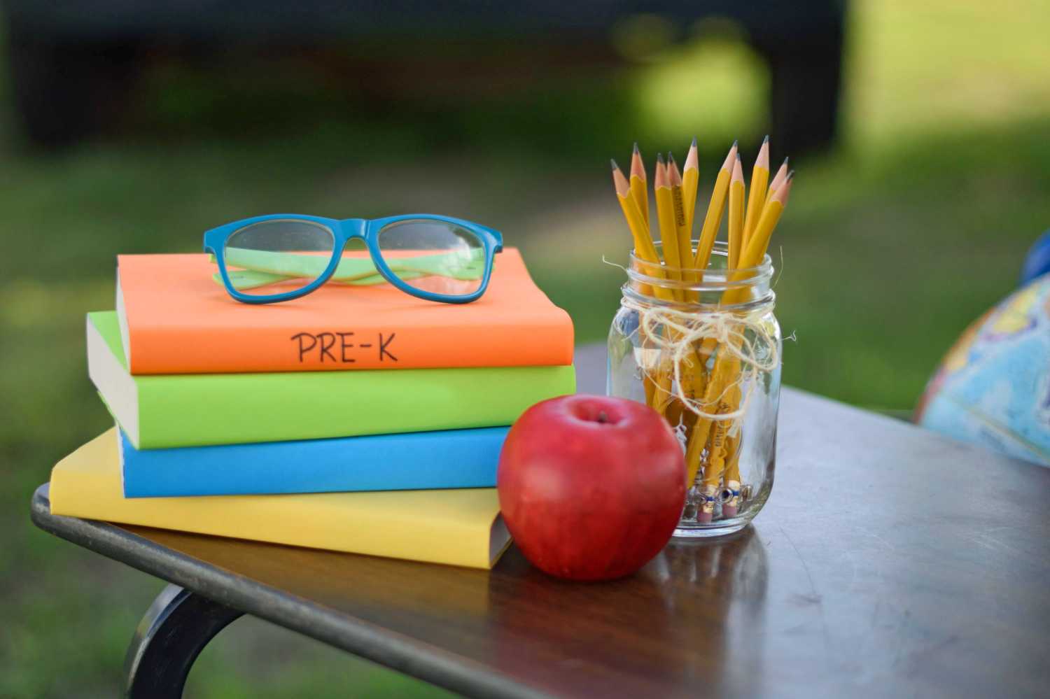 A stack of books on a table with pre-K written on the spine of one book and glasses sitting on top of the stack. Also, an apple and a glass of pencils sit on the table.