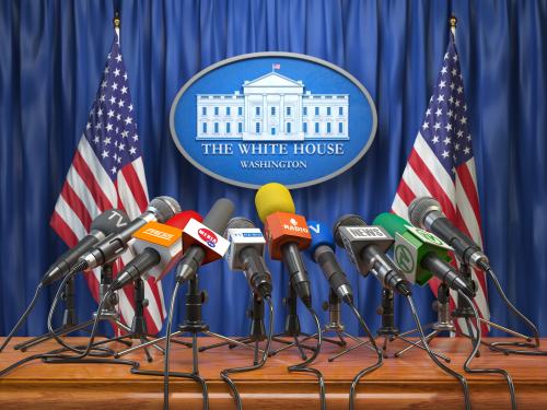 Press conference stand in the U.S. White House. Multiple microphones in front of stand and American flags in background.