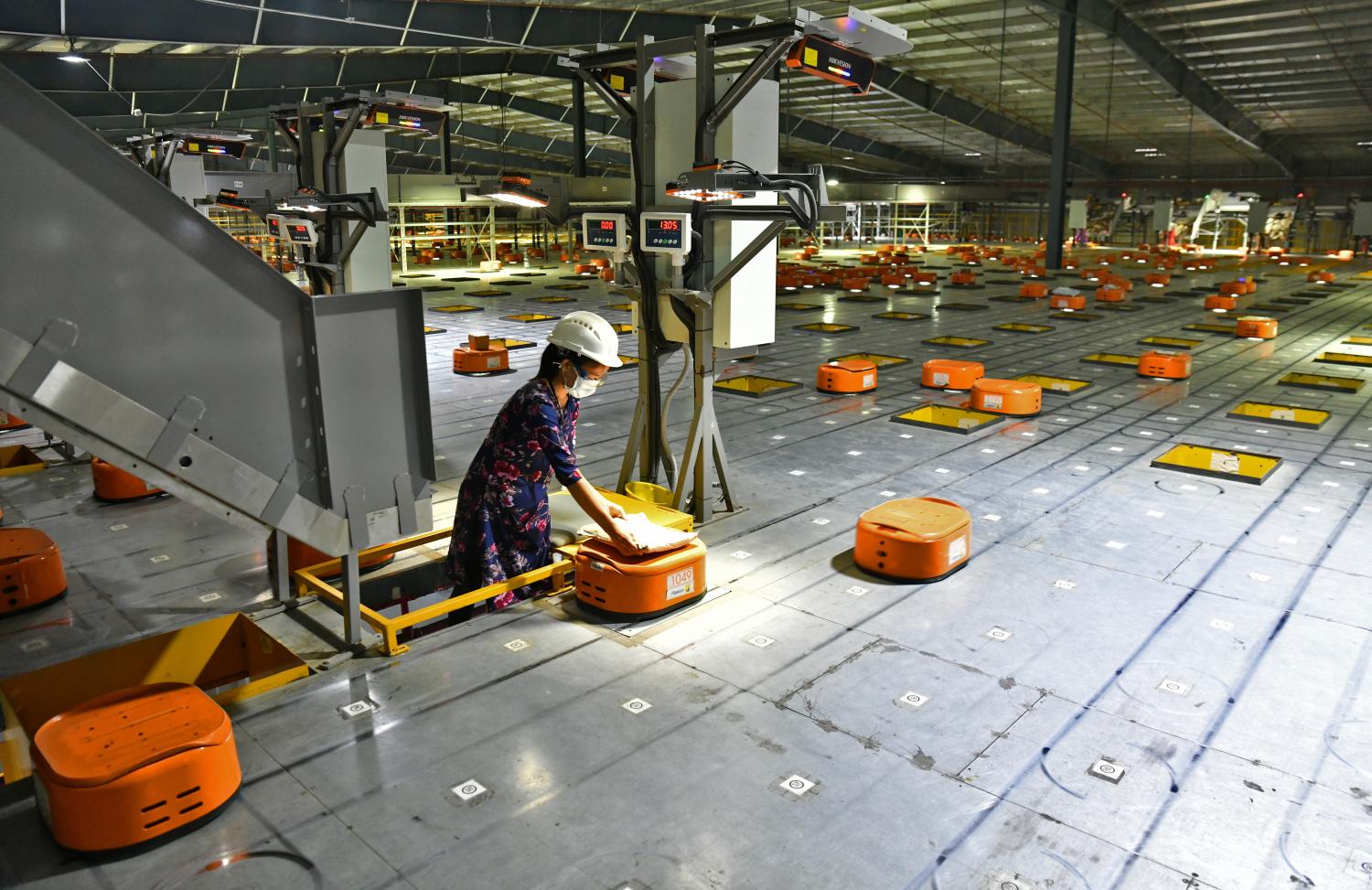 A worker stands in a large warehouse space, using small automated vehicles to sort items.