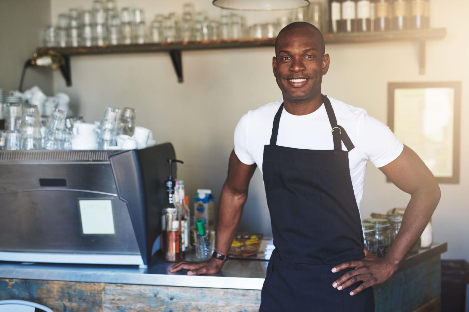 Young, Black male coffee house employee with white shirt and black apron at counter