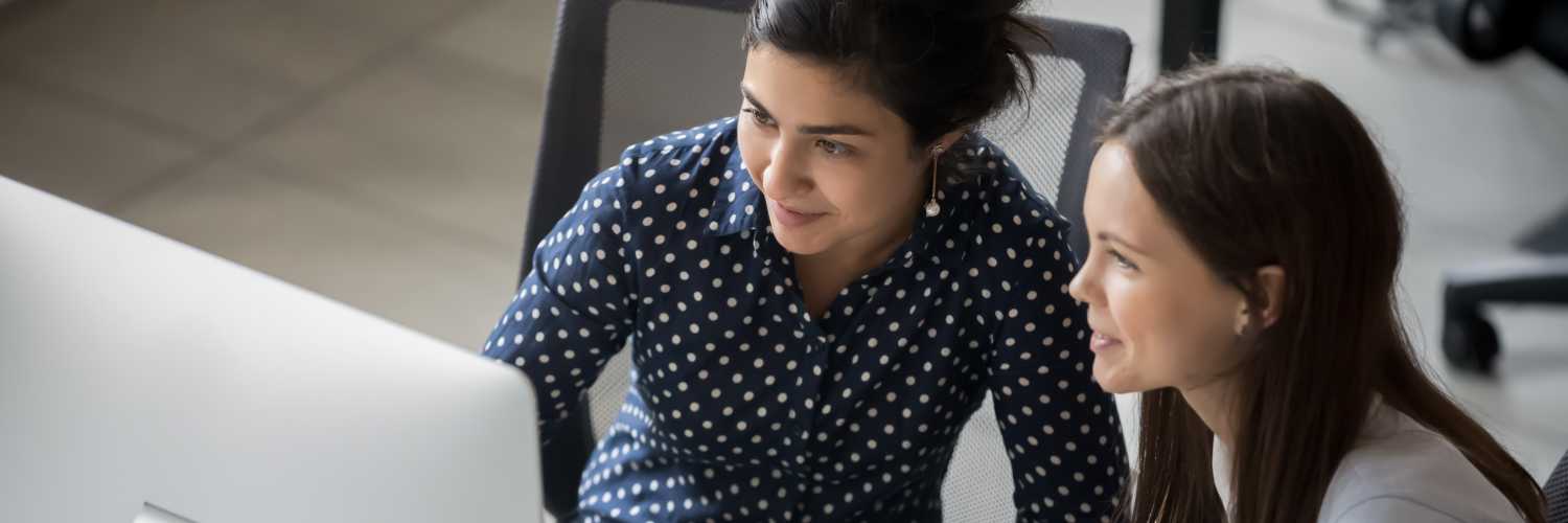 Multi ethnic female colleagues sit at workplace working together looks at pc screen use corporate program