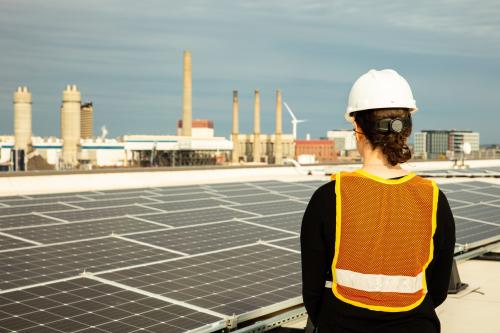 Female solar installer inspecting a commercial rooftop solar system in Charlestown, Boston, Massachusetts, U.S. with a wind turbine and a natural gas electric generation facility in the background.